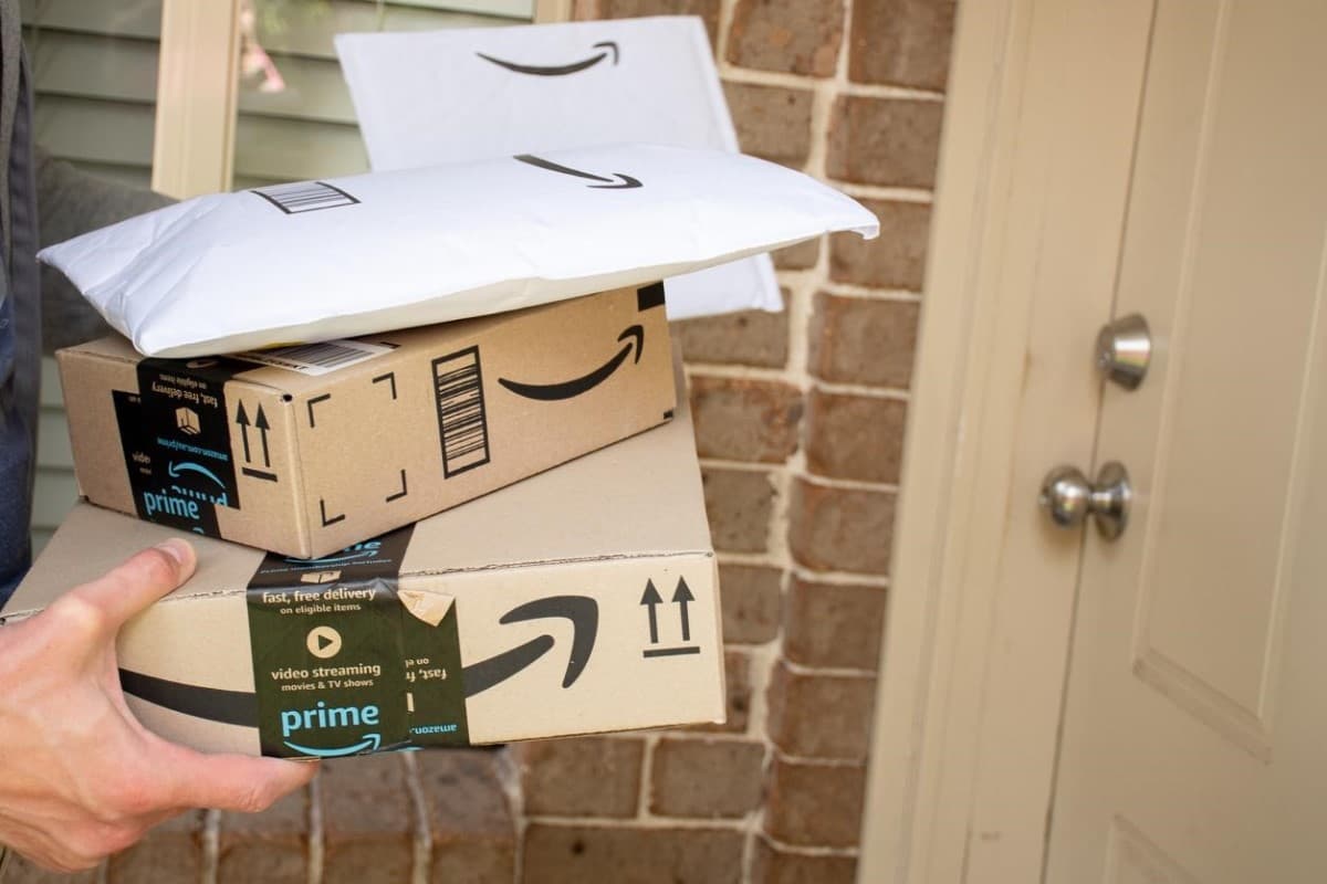 Un hombre con sobres y cajas de Amazon frente a una puerta. Aprende cómo comprar acciones de Amazon.